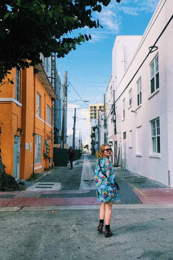 A passeggio per Miami Beach in una splendida giornata soleggiata, indossando una camicia azzurra in stile hawaiano che richiama il cielo limpido. Sullo sfondo, i vivaci palazzi Art Déco aggiungono un tocco iconico e colorato al paesaggio.