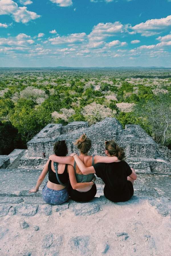 Foto di gruppo nello Yucatán per lo spring break messicano, sedute di spalle in cima alle antiche rovine di Calakmul, immersi nella vista mozzafiato della giungla rigogliosa di Campeche. Paesaggio verde e selvaggio che abbraccia l'orizzonte.