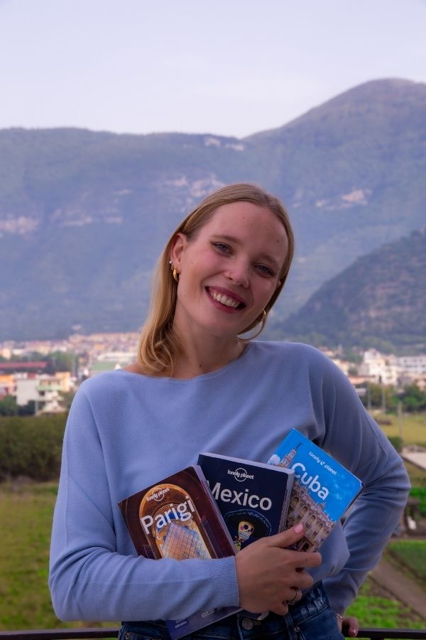 Io sorridente con capelli biondi lisci mentre tengo in braccio tre guide di viaggio Lonely Planet per Parigi, Messico e Cuba. Indosso un maglione celeste a maniche lunghe e mi trovo all'aperto, con uno sfondo di colline e montagne coperte di vegetazione. In lontananza si intravede una cittadina con edifici bassi e campi verdi, si tratta di Sarno. Il cielo è nuvoloso e l'atmosfera generale è tranquilla e serena.